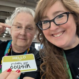 Katy Rowe-Schurwanz and Mags Gaulden smiling at RootsTech, holding a badge that reads &ldquo;Probably your cousin.&rdquo;