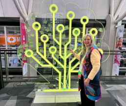 Roberta Estes standing in front of a neon RootsTech logo sign at the genealogy conference.